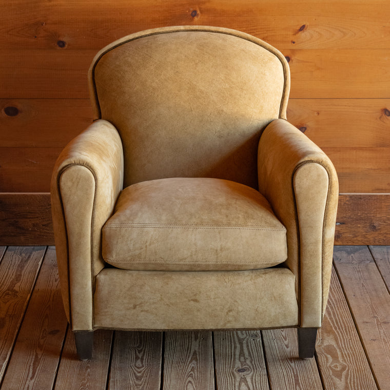 Angled back view of a tight back leather arm chair upholstered in a rustic high-contrast leather with coordinating welt on a wide-plank wood floor against a rustic wooden wall