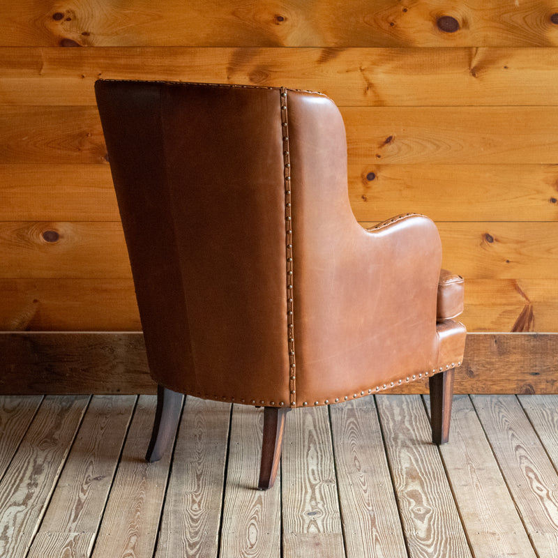 Angled back view of a brown leather wingback chair with nailhead trim against a wooden wall