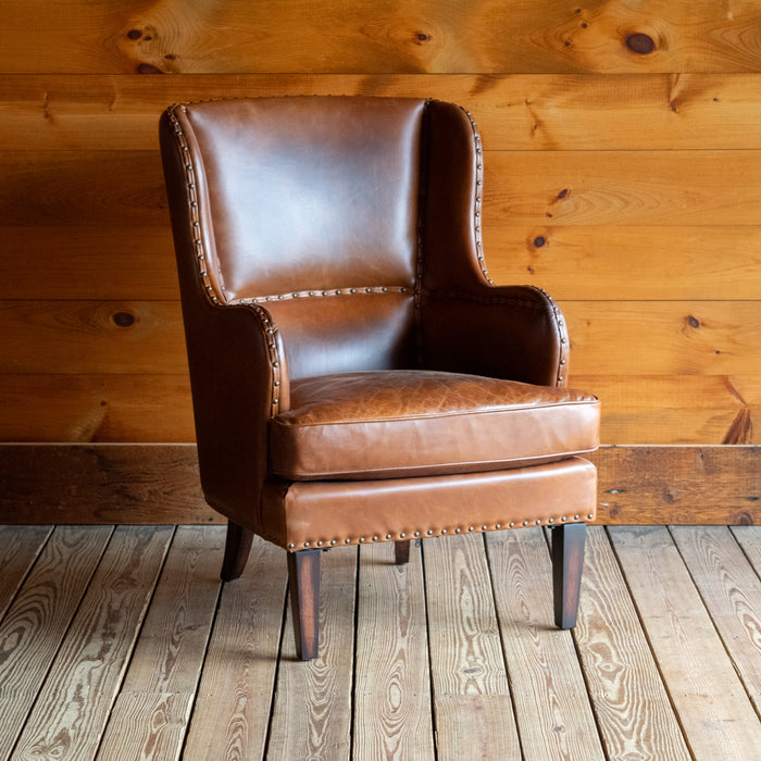 Angled front view of a brown leather wingback chair with nailhead trim against a wooden wall