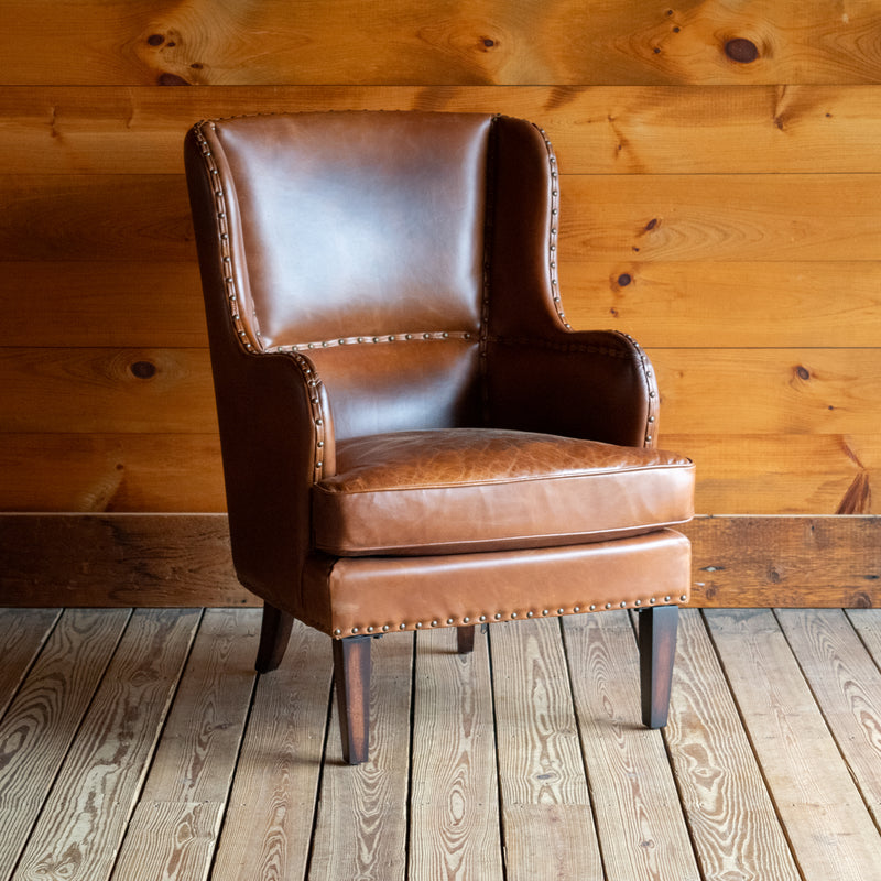 Angled front view of a brown leather wingback chair with nailhead trim against a wooden wall