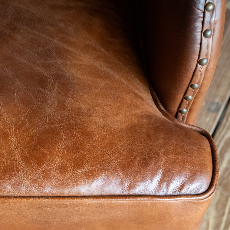 Angled back view of a brown leather wingback chair with nailhead trim against a wooden wall