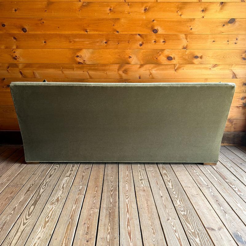 Back view of a tight back green velvet sofa with two matching throw pillows on a wooden floor against a wooden wall