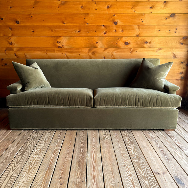 Back view of a tight back green velvet sofa with two matching throw pillows on a wooden floor against a wooden wall