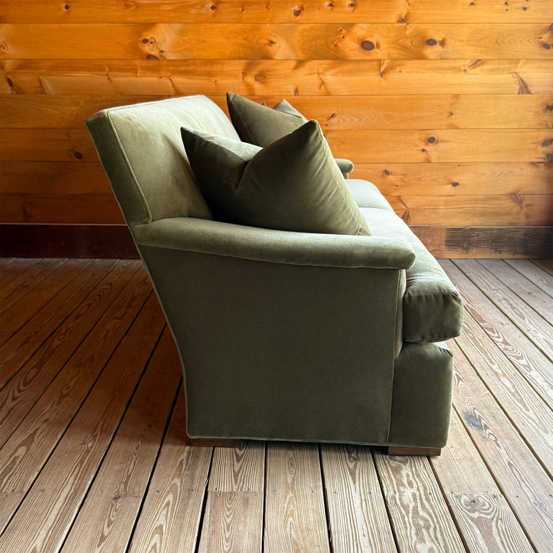 Profile view of a tight back green velvet sofa with two matching throw pillows on a wooden floor against a wooden wall