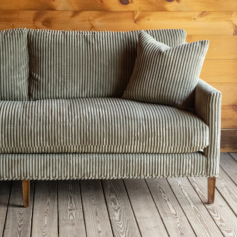 Front view close-up of a rustic sofa upholstered in a muted green corded velour fabric with a matching throw pillow against a wooden wall 