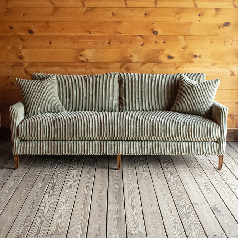 Back view of a rustic sofa upholstered in a muted green corded velour fabric against a wooden wall 