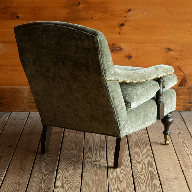 Angled back view of a muted green chenille open arm lounge chair with nailhead trim and turned wooden front legs on casters on a wooden floor against a wooden wall