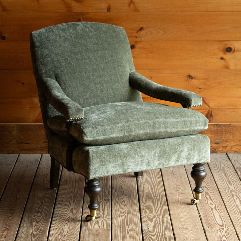 Angled front view of a muted green chenille open arm lounge chair with nailhead trim and turned wooden front legs on casters on a wooden floor against a wooden wall