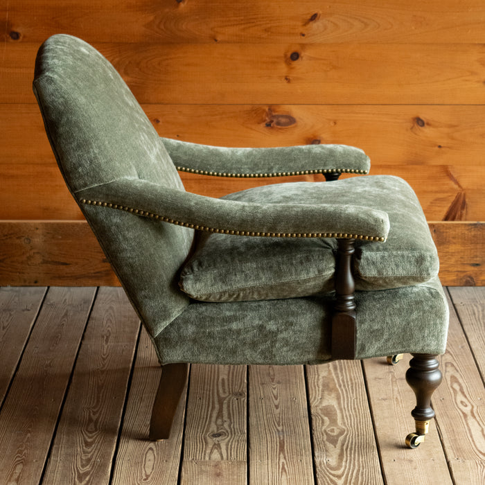 Profile view of a muted green chenille open arm lounge chair with nailhead trim and turned wooden front legs on casters on a wooden floor against a wooden wall