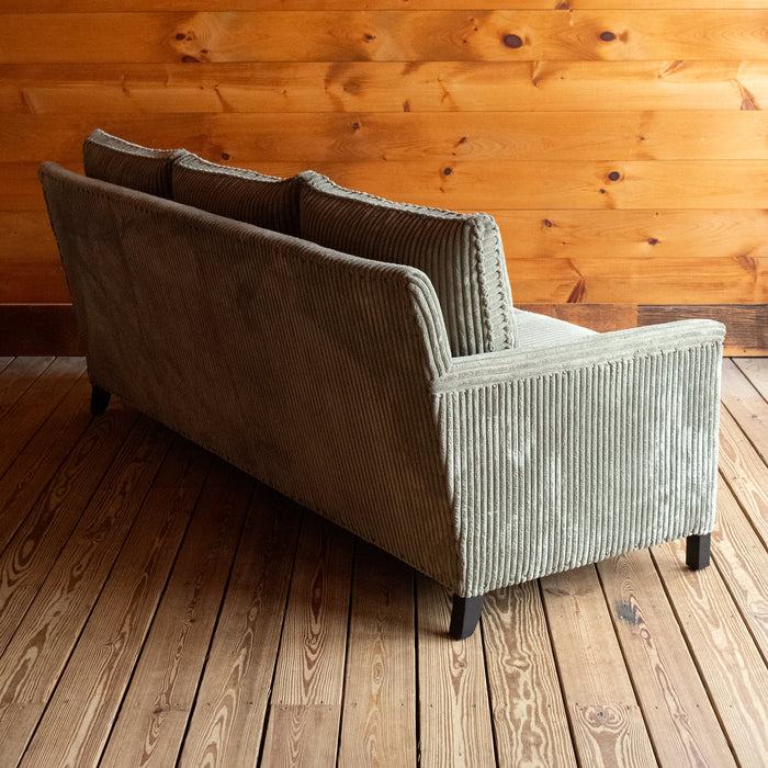 Angled back view of a three-seat sofa upholstered in a muted green corded velour fabric on a wooden floor against a wooden wall