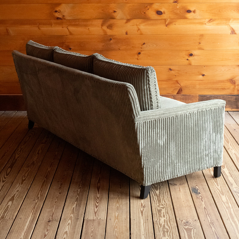 Angled back view of a three-seat sofa upholstered in a muted green corded velour fabric on a wooden floor against a wooden wall