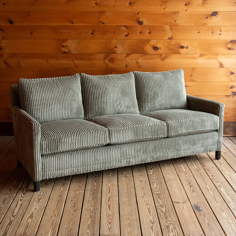 Angled front view of a three-seat sofa upholstered in a muted green corded velour fabric on a wooden floor against a wooden wall