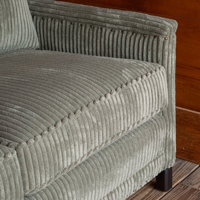 Angled back view of a three-seat sofa upholstered in a muted green corded velour fabric on a wooden floor against a wooden wall
