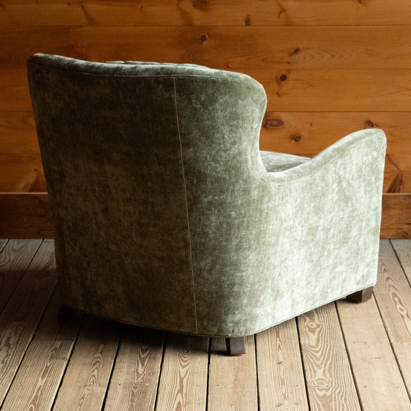 Angled back view of a deep, comfy wing-back chair upholstered in a mottled, muted green fabric on a wide-plank wooden floor against a rustic wooden wall