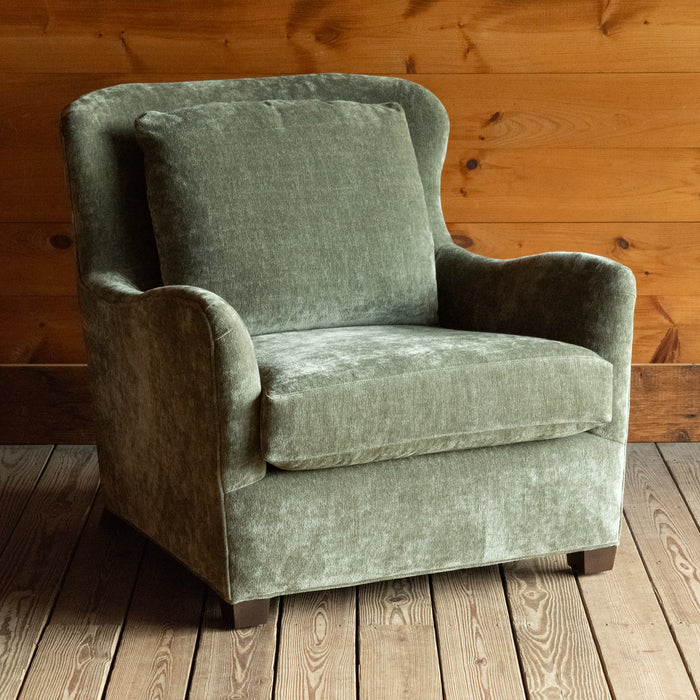 Angled front view of a deep, comfy wing-back chair upholstered in a mottled, muted green fabric on a wide-plank wooden floor against a rustic wooden wall