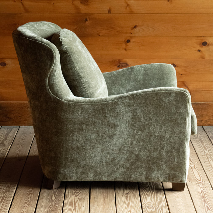Profile view of a deep, comfy wing-back chair upholstered in a mottled, muted green fabric on a wide-plank wooden floor against a rustic wooden wall