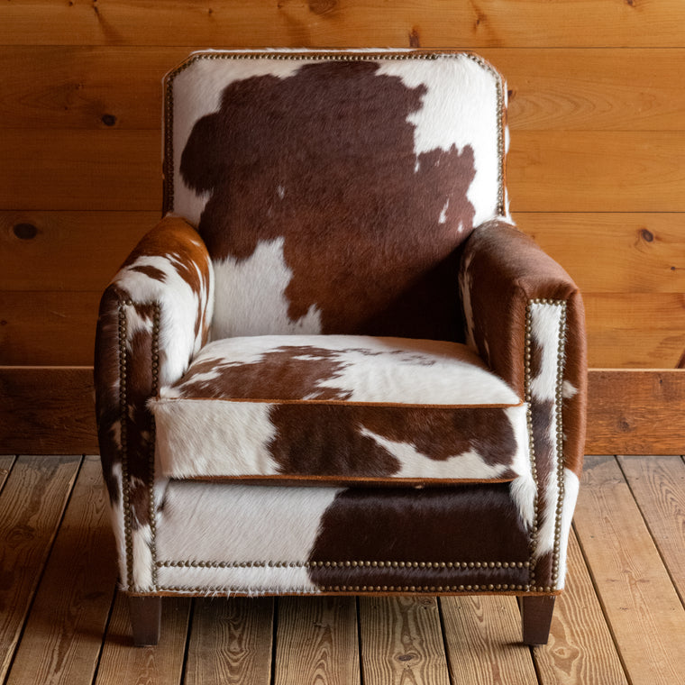 Angled back view of a rustic club chair upholstered in natural, brown and white cowhide leather on a wide-plank wooden floor against a rustic wooden wall
