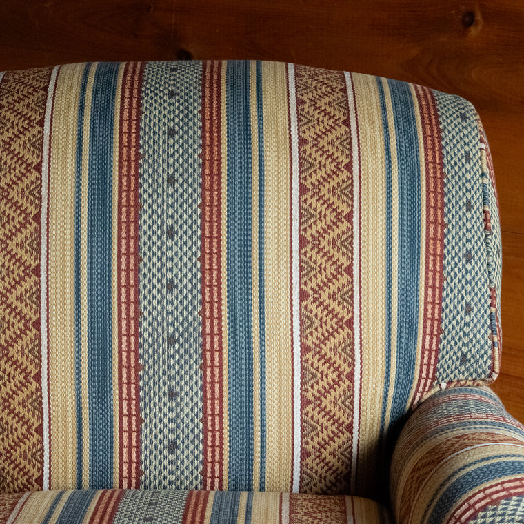 Angled back view of an oversized armchair upholstered in a rustic striped fabric, on a wide-plank wooden floor against a rustic wooden wall