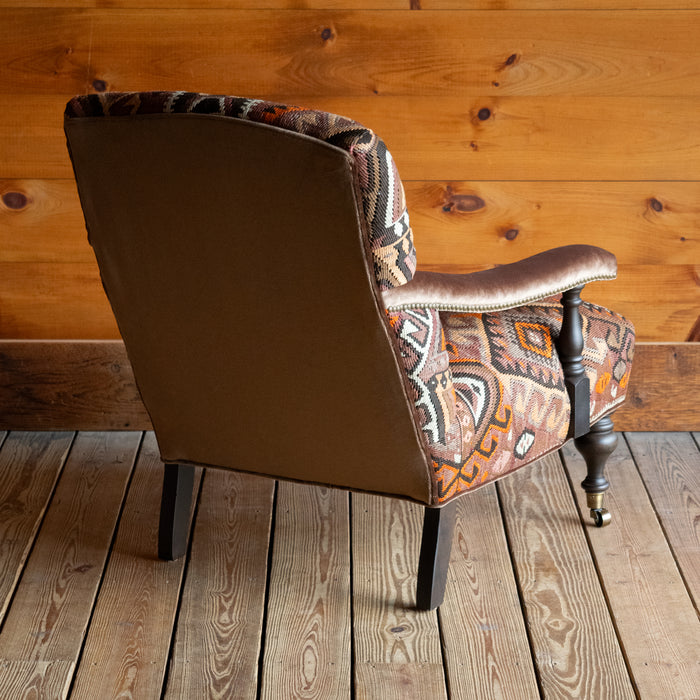 Angled back view of an armchair with brown, beige, and orange geometric patterned rug upholstery set on a wide-plank wooden floor against a rustic wooden wall