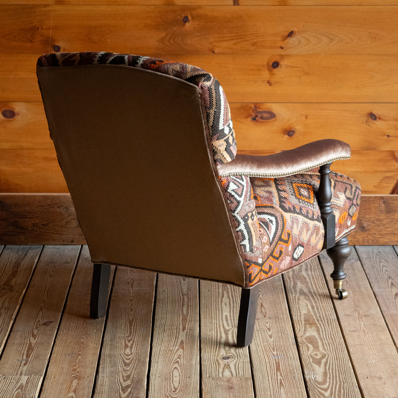 Angled back view of an armchair with brown, beige, and orange geometric patterned rug upholstery set on a wide-plank wooden floor against a rustic wooden wall