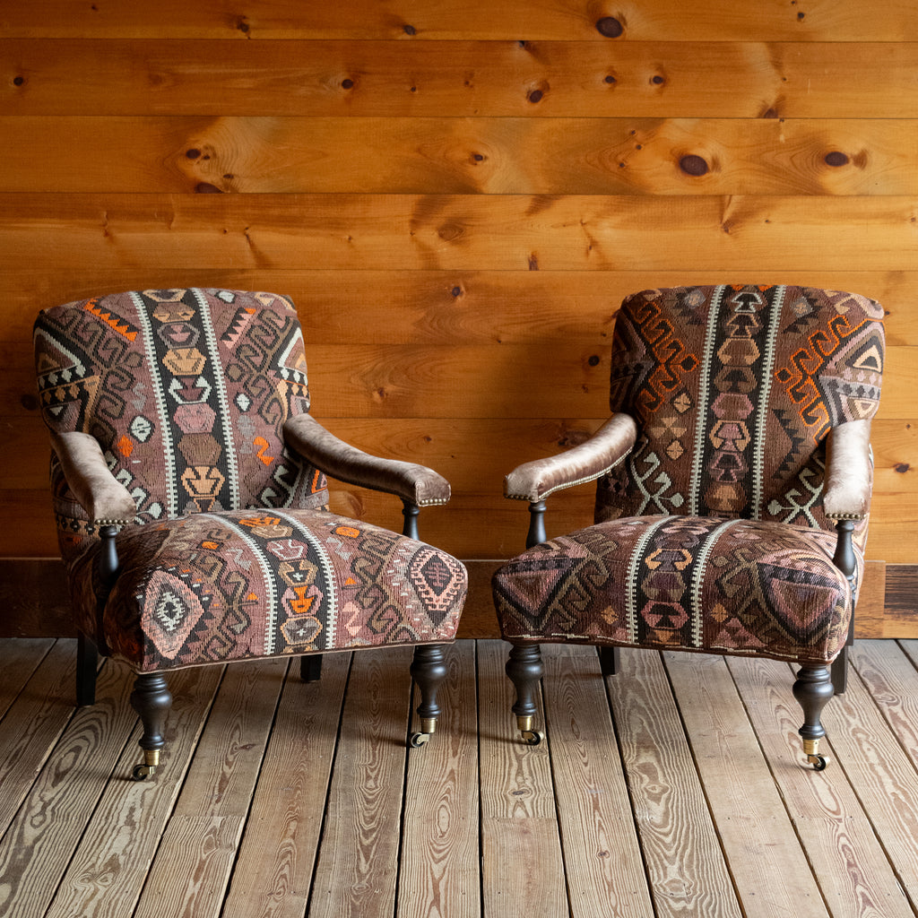 Two armchairs with brown, beige, and orange geometric patterned rug upholstery set on a wide-plank wooden floor against a rustic wooden wall
