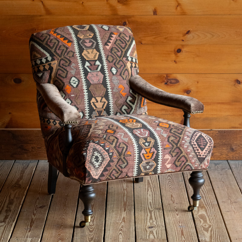 Angled front view of an armchair with brown, beige, and orange geometric patterned rug upholstery set on a wide-plank wooden floor against a rustic wooden wall