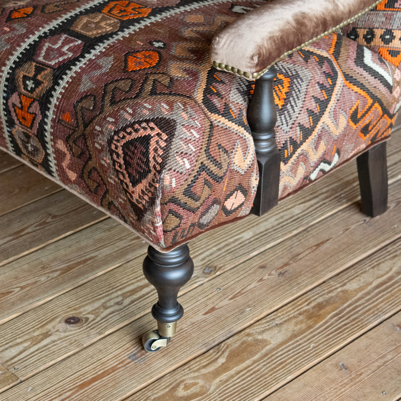 Close-up of the arm and leg of an armchair with brown, beige, and orange geometric patterned rug upholstery on a wide-plank wooden floor 