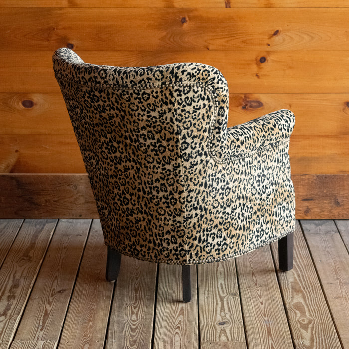 Angled back view of a petite arm chair upholstered in a jaguar print fabric on a wooden floor against a wooden wall