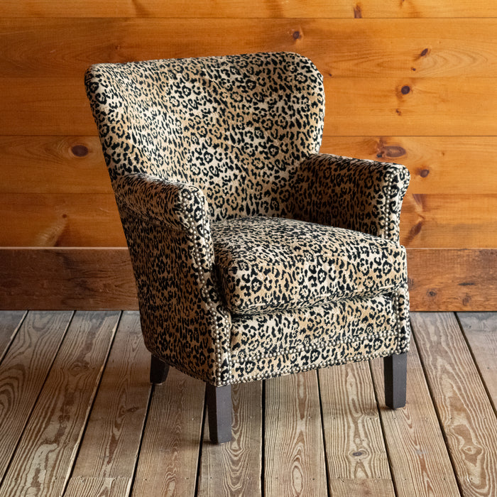 Angled front view of a petite arm chair upholstered in a jaguar print fabric on a wooden floor against a wooden wall