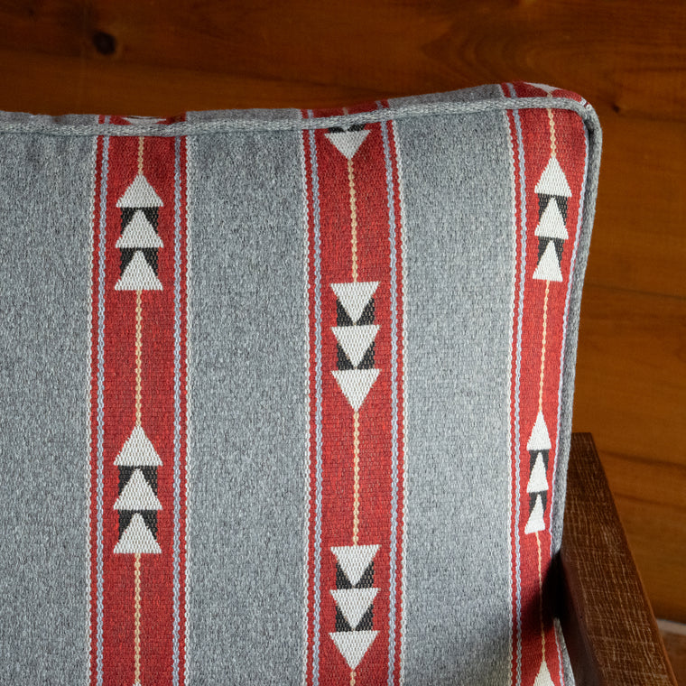 Close-up of the back and cushion of a rustic chair with light gray, red, and white geometric striped cushions on a reclaimed red oak frame, on a wide-plank wood floor against a rustic wooden wall