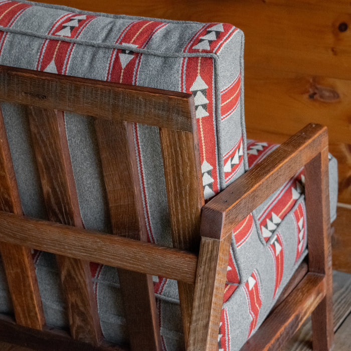 Close-up of the back and cushion of a rustic chair with light gray, red, and white geometric striped cushions on a reclaimed red oak frame, on a wide-plank wood floor against a rustic wooden wall