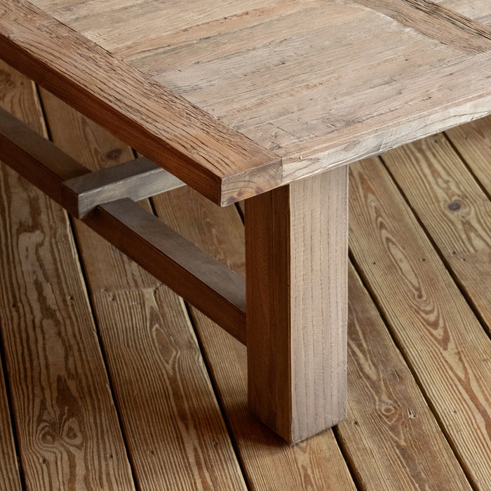 Close-up of the tabletop corner, angled leg, and stretchers, of a large, rectangular reclaimed oak coffee table on a wide-plank wooden floor