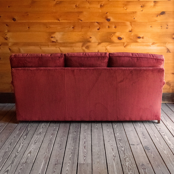 Back view of a red corduroy velvet sofa with English rolled arms, turned legs, and matching throw pillows on a wooden floor against a wooden wall 