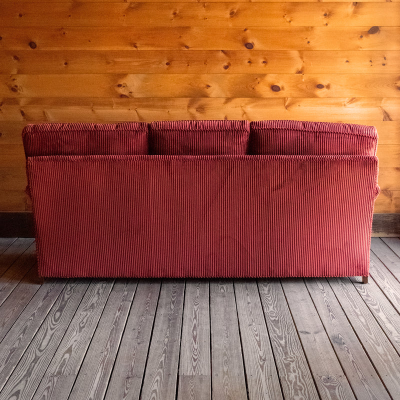 Back view of a red corduroy velvet sofa with English rolled arms, turned legs, and matching throw pillows on a wooden floor against a wooden wall 
