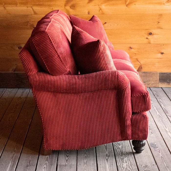 Profile view of a red corduroy velvet sofa with English rolled arms, turned legs, and matching throw pillows on a wooden floor against a wooden wall 