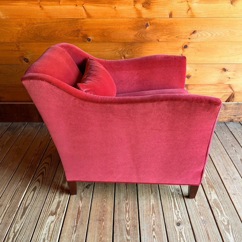 Profile view of a red velvet armchair with a lumbar pillow against a wooden wall