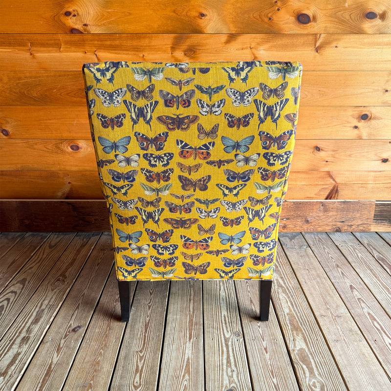 Back view of an armchair upholstered in a yellow-gold fabric with an allover butterfly pattern against a wooden wall