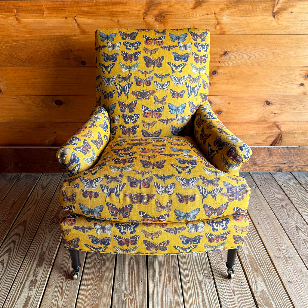 Front view of an armchair upholstered in a yellow-gold fabric with an allover butterfly pattern against a wooden wall