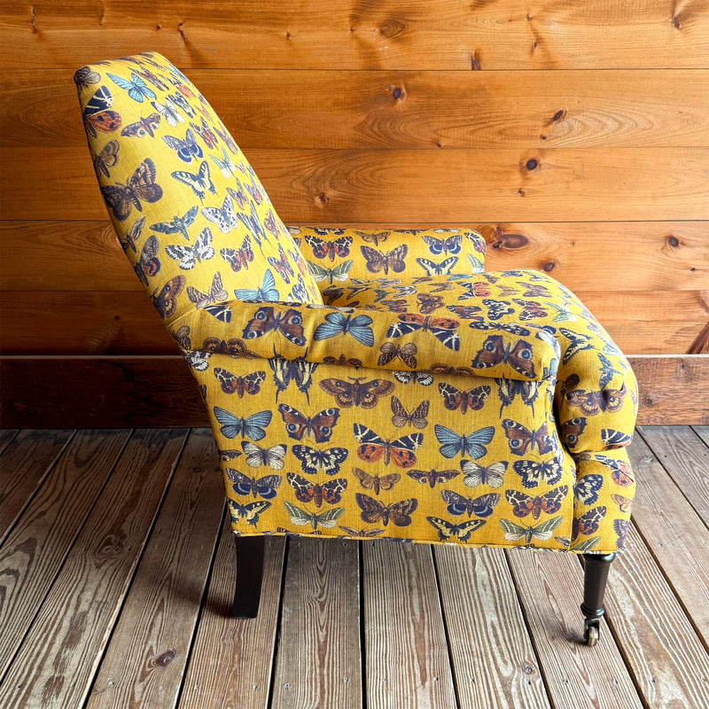 Profile view of an armchair upholstered in a yellow-gold fabric with an allover butterfly pattern against a wooden wall