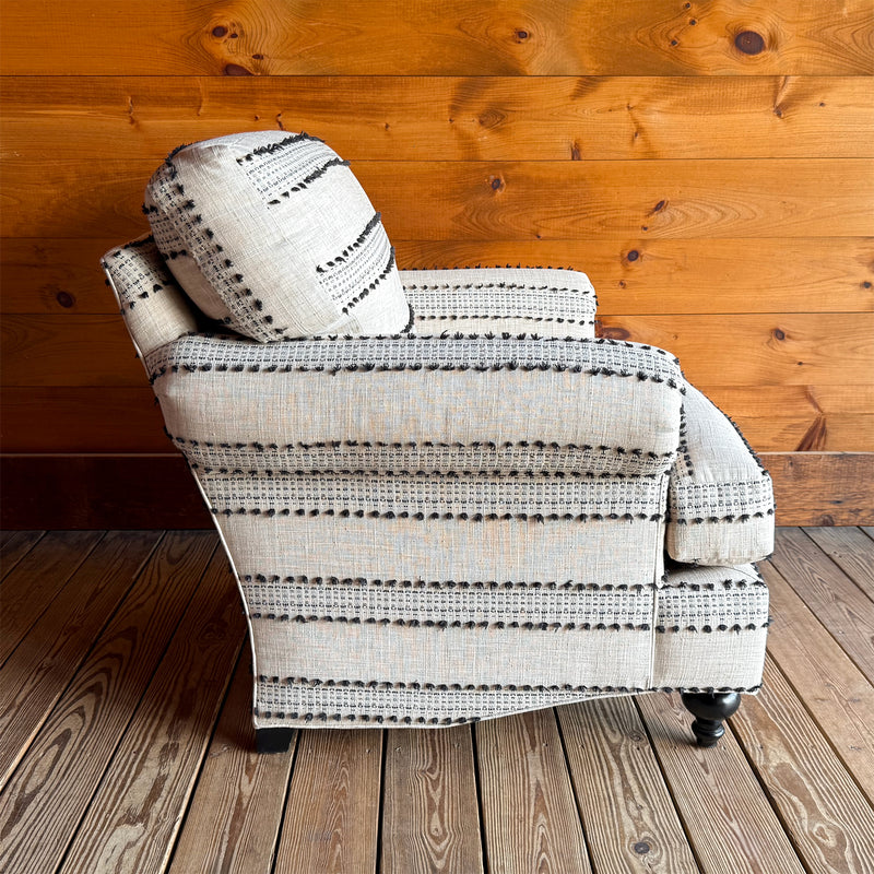 Profile view of ivory and black striped roll arm chair with turned front legs against wooden wall.