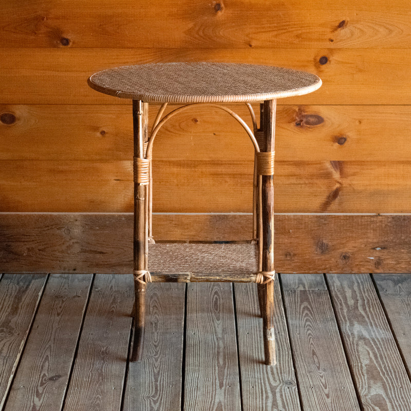 Front view of a bamboo side table with a round woven top against a wooden wall.