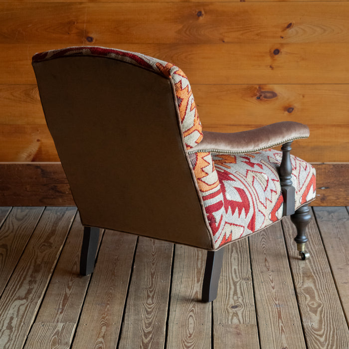 Angled back view of an armchair upholstered in a rug fabric featuring a large-scale geometric pattern in crimson, rust, and light blue-gray, set on a wide-plank wooden floor against a rustic wooden wall