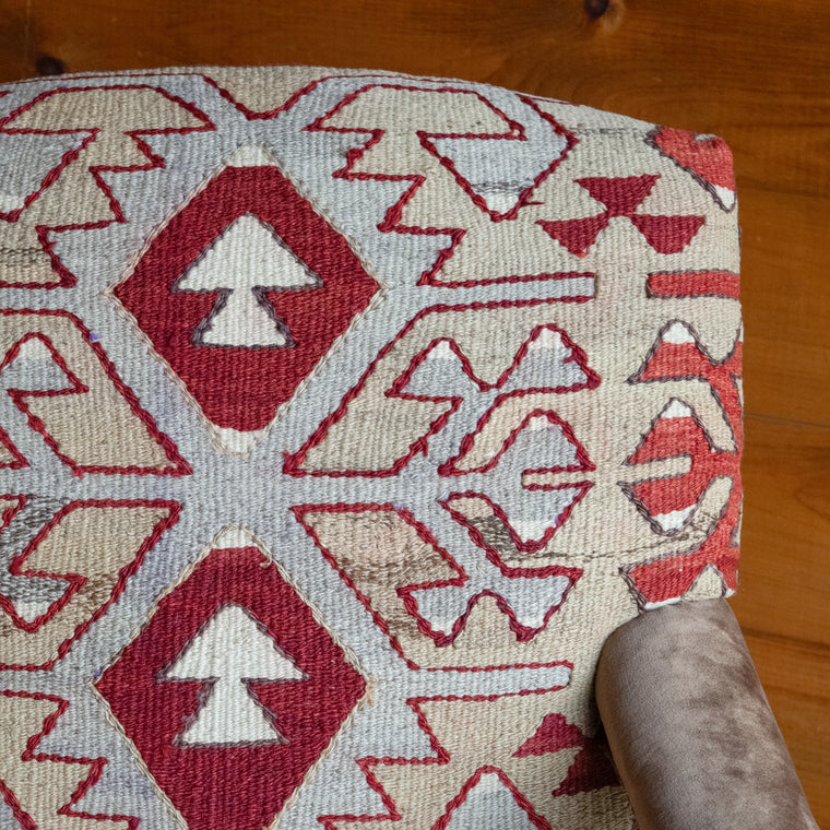Angled back view of an armchair upholstered in a rug fabric featuring a large-scale geometric pattern in crimson, rust, and light blue-gray, set on a wide-plank wooden floor against a rustic wooden wall