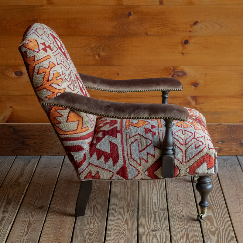 Profile view of an armchair upholstered in a rug fabric featuring a large-scale geometric pattern in crimson, rust, and light blue-gray, set on a wide-plank wooden floor against a rustic wooden wall