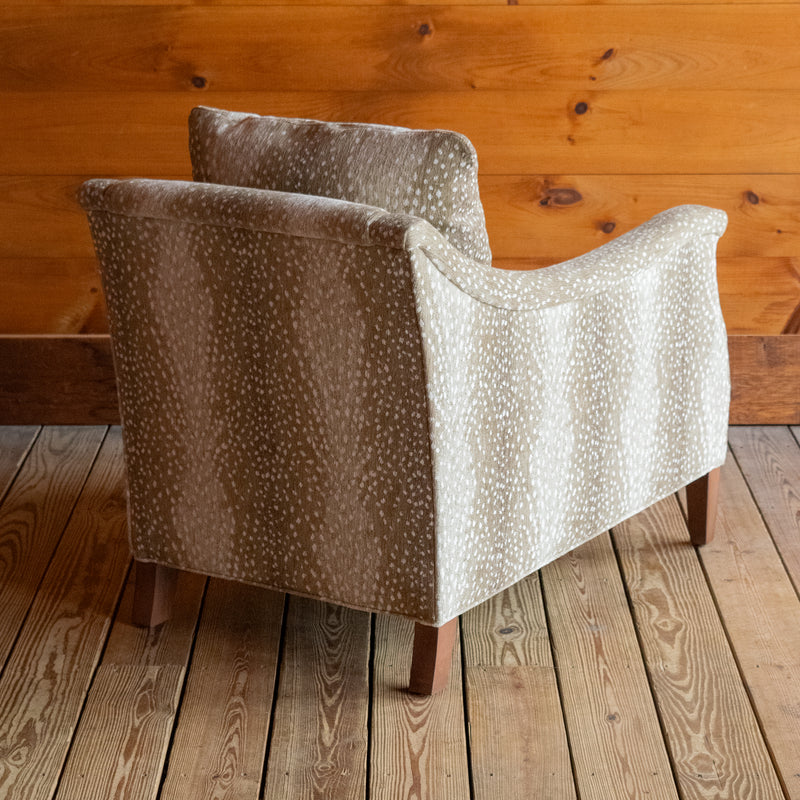 Angled back view of a sloped arm chair upholstered in a spotted antelope-inspired fabric with tapered hardwood legs on a wooden floor against a wooden wall