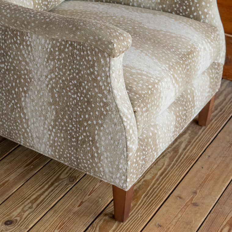 Angled back view of a sloped arm chair upholstered in a spotted antelope-inspired fabric with tapered hardwood legs on a wooden floor against a wooden wall