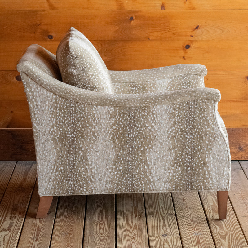 Profile view of a sloped arm chair upholstered in a spotted antelope-inspired fabric with tapered hardwood legs on a wooden floor against a wooden wall