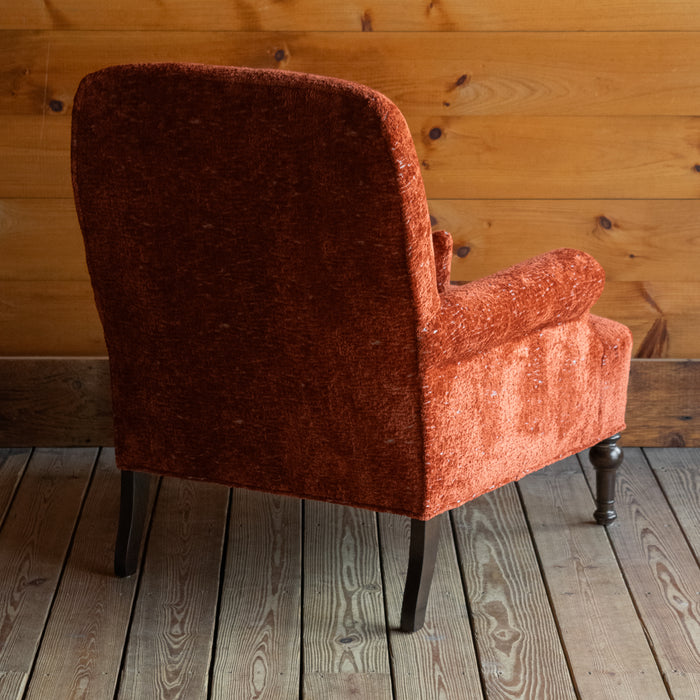 Angled back view of a tomato-red armchair with a tight seat and back on a wide-plank wood floor against a rustic wooden wall