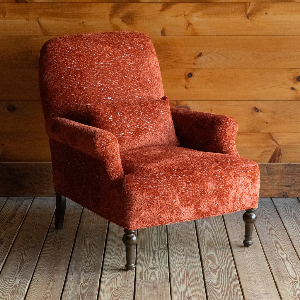Angled front view of a tomato-red armchair with a tight seat and back on a wide-plank wood floor against a rustic wooden wall