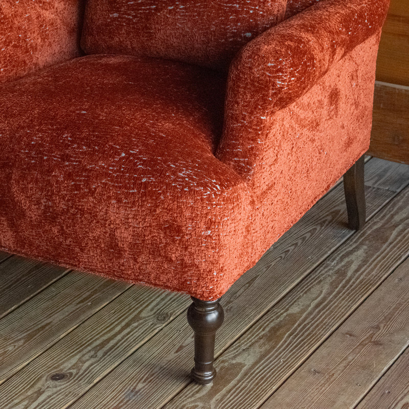 Close-up of the arm and leg of a tomato-red armchair with a tight seat and back on a wide-plank wooden floor